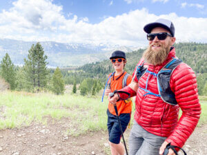 Two hikers stand together outdoors on a trail, with mountains and open landscape in the background.