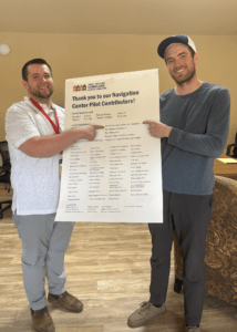 Justin Jones, Navigation Center Director, and Josh Landheer, Whole Person Care Coordinator, smile while holding up a printed document together.