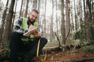 A man in a reflective safety vest kneels in a forest, taking notes on a clipboard near a fallen log.