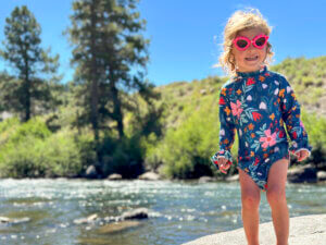 A young child in sunglasses stands along a rocky riverbank on a sunny day, with trees and hills in the background.